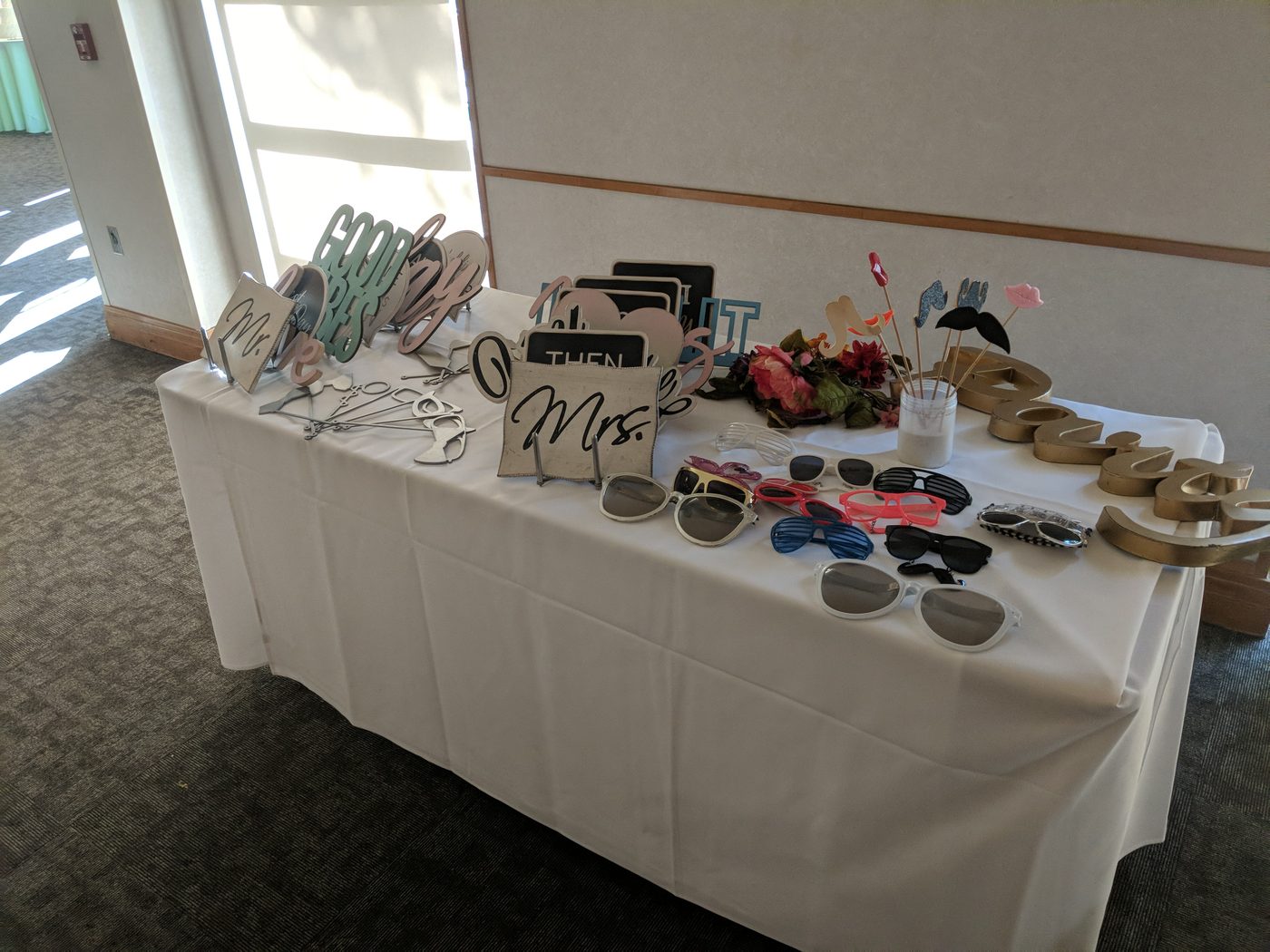Photobooth prop table with a 'Mrs' sign and florals at a sunlit venue
