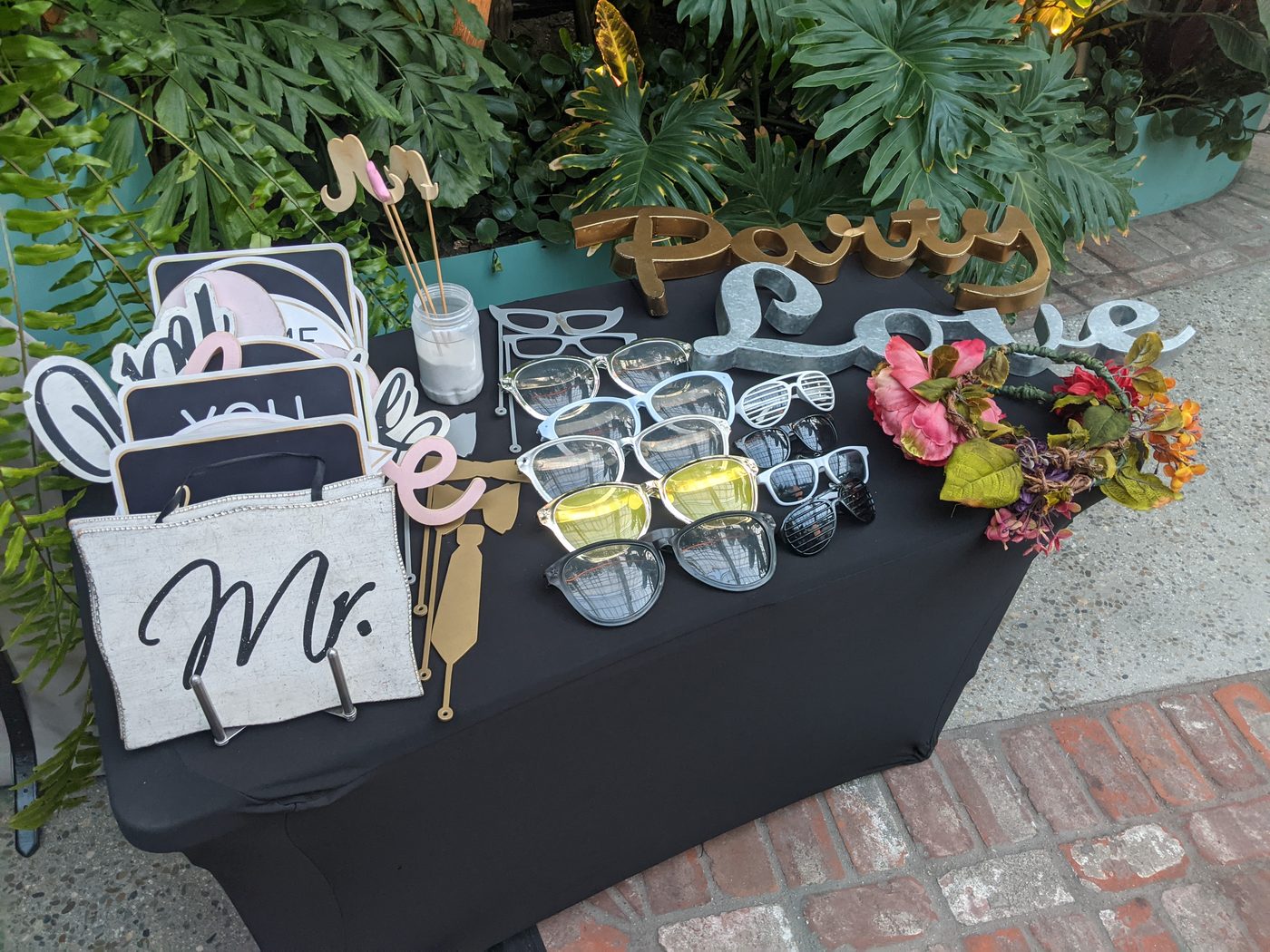Photobooth prop table with a 'Mr.' sign, floral crown, and sunglasses among tropical greenery