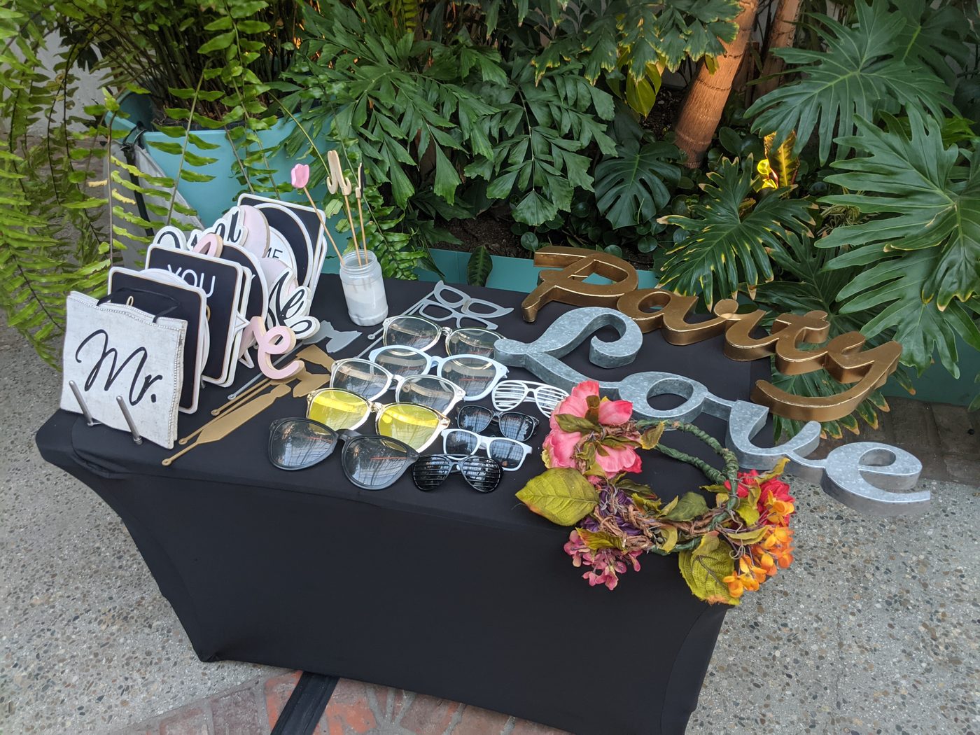 Photobooth prop table with 'Party Love' sign cutouts and sunglasses among tropical greenery